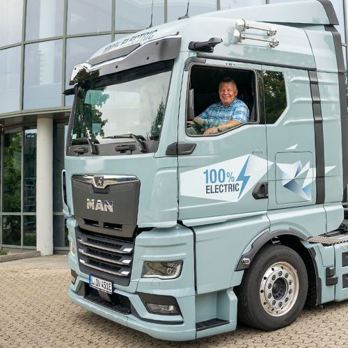 Person sitzt am Steuer eines hellblauen Elektro Lkw vor einem modernen Glasgebäude, dem Hauptsitz von Schnellecke Logistics in Wolfsburg.