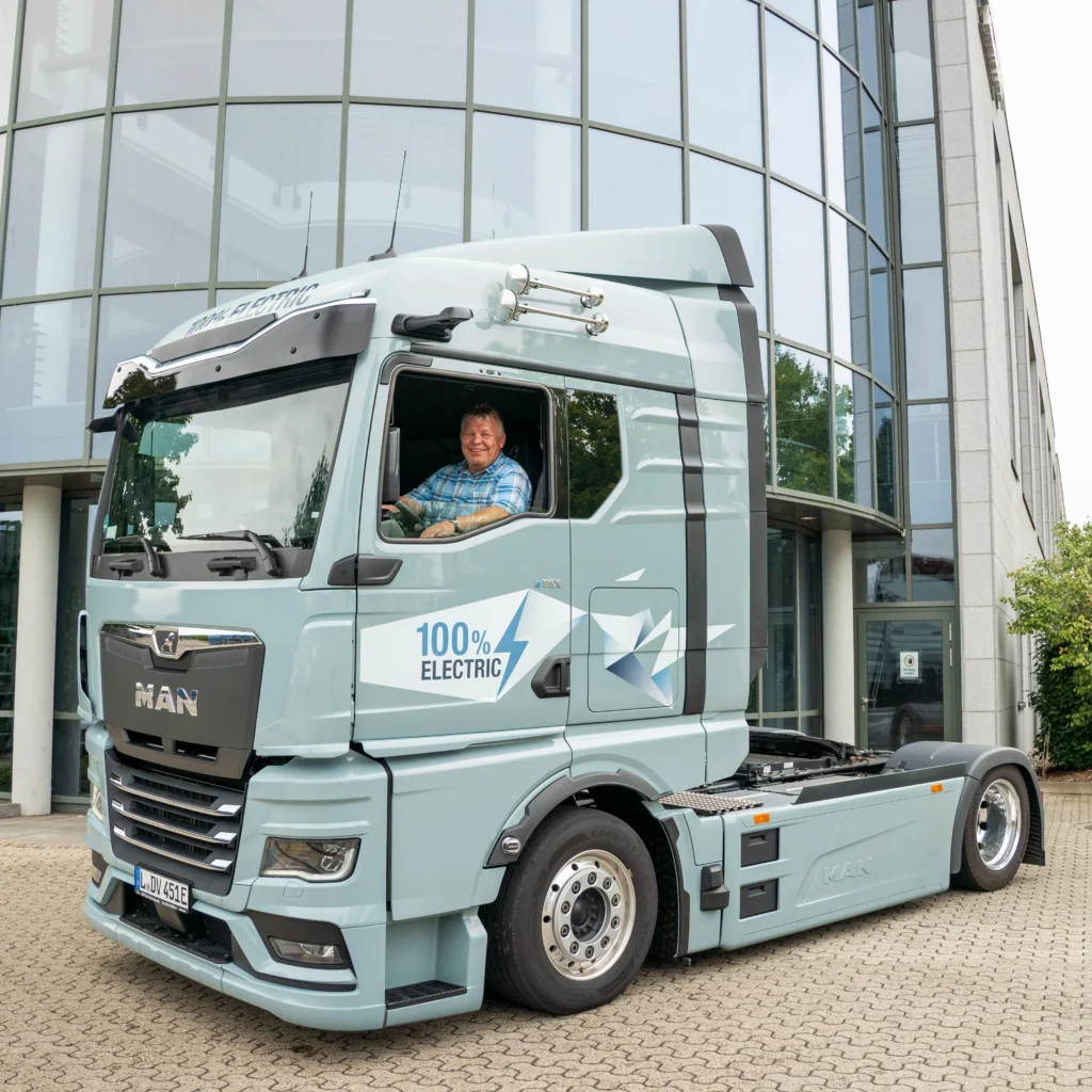 Person sitzt am Steuer eines hellblauen Elektro Lkw vor einem modernen Glasgebäude, dem Hauptsitz von Schnellecke Logistics in Wolfsburg.